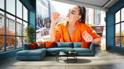 Stylish young woman sits in a street cafe and eats pizza. Beautiful brunette in a bright orange jacket. Delicious quick lunch. Wall mural