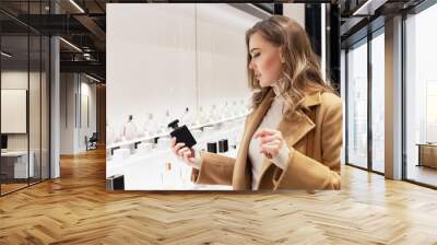 Young woman chooses perfume in the store. Smiling beautiful blonde in a beige coat. Individual scent and expensive cosmetics. Wall mural