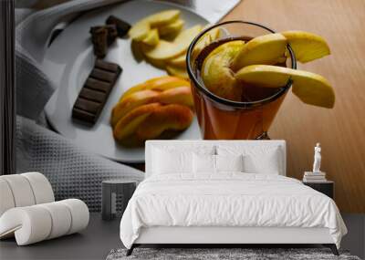 Apple tea in a transparent glass with a handle, the drink is decorated with slices of fresh apple with a yellow peel, next to the plate of sliced fruit and chocolate, everything is on the table on a w Wall mural