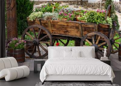 Lush floral arrangement of plants blooming with different colors and green leaves in a brown wooden cart with wheels. Mobile decorative table in natural rustic style Wall mural