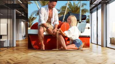 handsome man and blond woman enjoying their vacation on a sunny sand beach sitting on a boat, they are happy on summer day. a couple in love having fun on an island, beautiful family likes o travel Wall mural