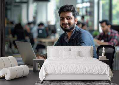 An intelligent and handsome Indian office worker sits at his desk working on a laptop. In the background, a diverse team of young professionals are at work. Wall mural