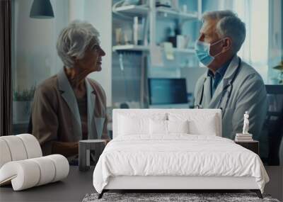 An older family doctor talks with a female senior patient in a health clinic while both wear masks. The doctor is seated behind a desk in a hospital office wearing a lab coat. Wall mural