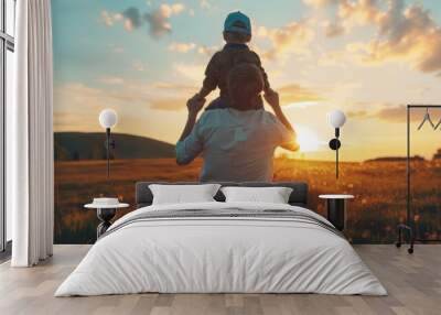 Father's day. View of a little boy seated on his father's shoulders holding hands and looking into the distance while enjoying the sunset. Father walking with son outdoors. Wall mural