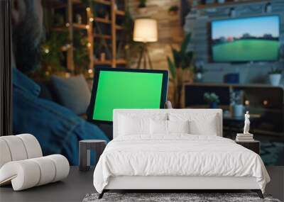 Sitting at the desk, man watching videos on a green mock-up screen of a digital tablet computer. The background shows a cozy living room in the background as the man watches videos. Wall mural