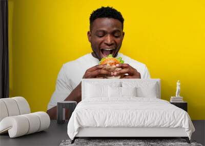 african american man in white t-shirt bites big burger on yellow isolated background, young guy eats fast food Wall mural