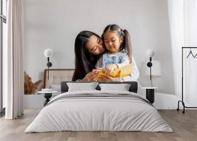 asian family sitting on bed and reading book at home, korean woman mother teaching little daughter to read book Wall mural