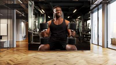 athletic african american man is training in dark gym and smiling, sports guy is sitting and holding heavy dumbbells Wall mural