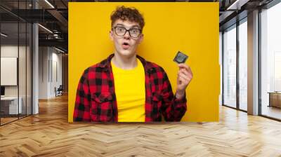 curly guy holds a condom on a yellow background and is surprised, a student shows contraceptive, the concept of safe sex Wall mural