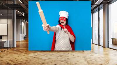 girl chef in superman costume holds kitchen item on blue background, woman housewife in superhero mask points forward Wall mural