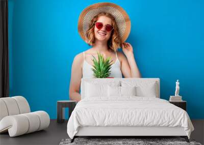 girl in a sun hat and glasses holds a pineapple on a blue isolated background and smiles, woman tourist on vacation, summer concept Wall mural