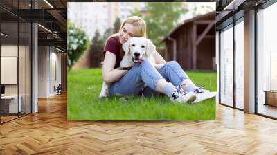 girl with a puppy of a retriever sits on a green grass in the spring on the street, a woman plays with a dog Wall mural