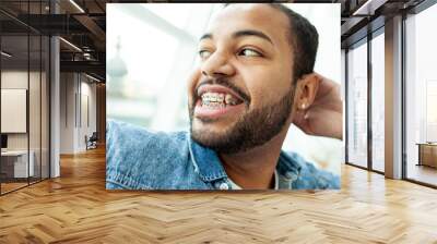 happy african american man with braces laughs and looks out the window in a white cafe, relaxed man in a denim shirt dreams and smiles Wall mural