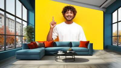 I have an idea. young indian man in white t-shirt raises his finger and points up on yellow isolated background, curly guy knows solution and idea Wall mural