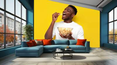 joyful african american man holding plate of chips and biting snack on yellow isolated background Wall mural