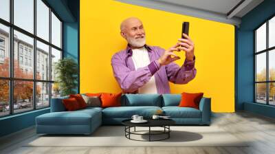 old bald grandfather with gray beard uses smartphone and selects online on yellow isolated background Wall mural