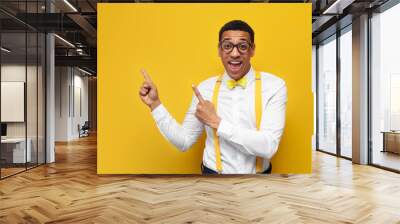 young african american guy in festive outfit and glasses shows with his hands on copy space on yellow background Wall mural