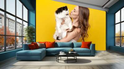 young cute girl kisses black and white cat on yellow isolated background and smiles, woman with pet Wall mural