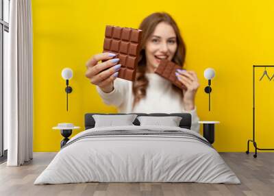 young girl eats and bites big chocolate bar and offers piece to you on yellow isolated background Wall mural