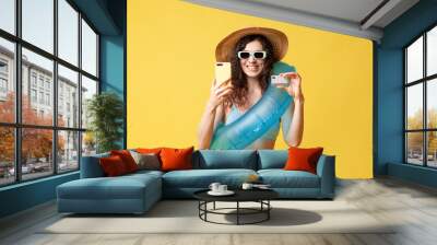 young girl in a blue swimsuit with a swimming inflatable ring holds smartphone and bank card on a yellow background Wall mural