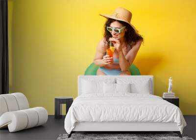 young girl in blue swimsuit sits on inflatable swimming ring holds orange alcoholic cocktail on yellow background Wall mural