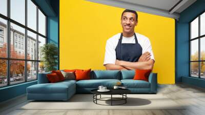 young guy afro american waiter in apron stands with his arms crossed on yellow isolated background, barista worker Wall mural