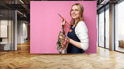 Young happy girl artist holds drawing equipment and draws with a brush on a pink background, the work of the artist Wall mural