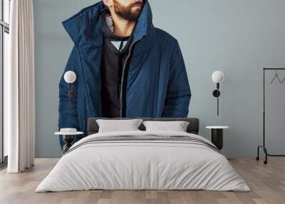 The guy model on a gray background is dressed in a blue down jacket. Hands in the pockets of a warm jacket. A guy with a beard and warmly dressed. Stylish warm clothes Wall mural