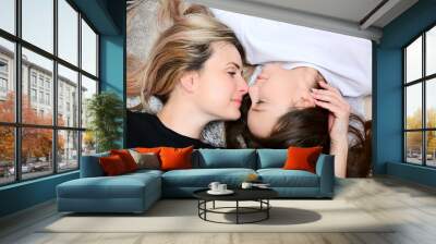 A beautiful blonde mother and a cute dark-haired daughter lie on the pillows and kiss each other. View from above. Happy motherhood Wall mural