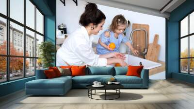 A beautiful young woman with her two year old daughter is washing fruit in the kitchen sink. Fresh bright fruits, apples, oranges, pears. Healthy food for children. The baby helps her mother. Wall mural