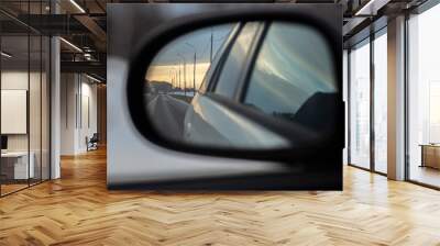 Reflection in the rearview mirror of a car. It displays a highway, lampposts along it, cars driving along the road, and in the background you can see the rays of the setting sun.  Wall mural