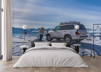 A man in a down jacket stands on the edge of a snowy plateau, leaning on an SUV. Footprints in the snow. In the distance - a mountain range, a valley. Blue sky. Altai Wall mural