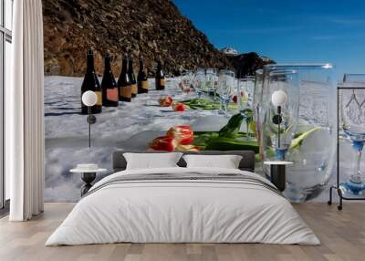 Early spring festival. Wine glasses and champagne bottles stand in a row. Bright red and yellow tulips lie on the white snow. Background - rock and blue sky. Wall mural