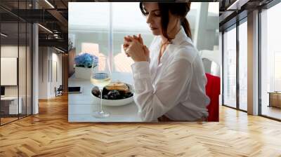 Adult beautiful woman sit in elite restaurant with a beautiful view of the sea, her order seafood and drink. The female person wearing white shirt. Girl chosses the italian wine and mussels. Wall mural