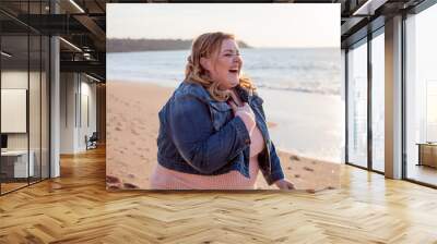 Beautiful overweight woman walking on the sandy beach. Plus size girl enjoy warmth sunset with romantic mood. Fat model dressed jeans jacket and pink knitted sweater Wall mural