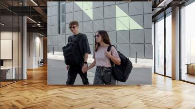 The modern couple of girl and boy teenagers dressed black t-shirt, jeans, sunglasses, watches and bagback walking in the street at the background grey wall Wall mural