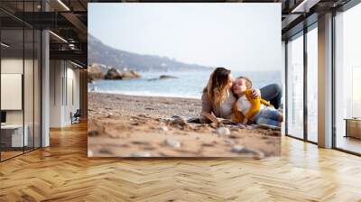 Woman read the book and relax in the blanket in the beach against stone while kid listen. Mother hugging with baby. Happy leisure to relax with family on the sea shore Wall mural