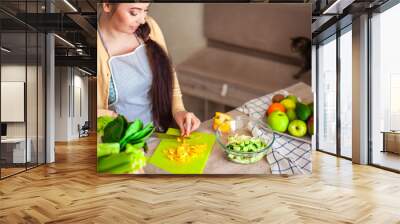 Young brunette woman making healthy food on the dinner table in the kitchen. Female cooking fresh vegan salad with green vegatable Wall mural