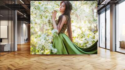 A beautiful young woman in a green dress with a large train among the blooming apple trees. Wall mural