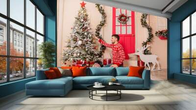 A young man in a red sweater with reindeer decorates a Christmas tree on the porch. Wall mural