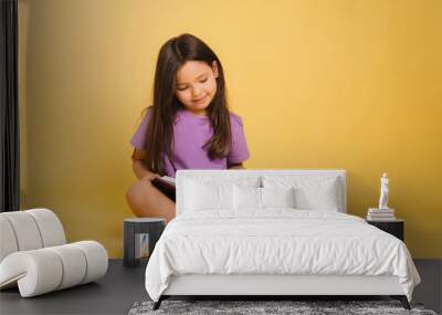 beautiful little girl in a purple t-shirt reads a book while sitting on a yellow background. Cute child learns lessons. Wall mural