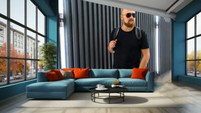 Handsome young stylish man with sunglasses and redhead beard in a black t-shirt poses near the gray wall Wall mural