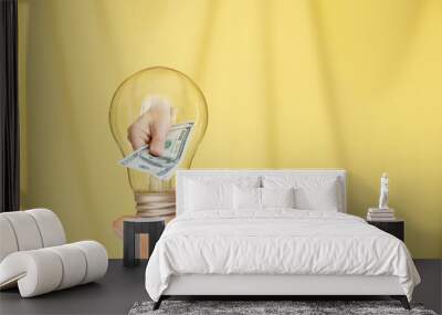 A woman's hand holds a light bulb and the second hand gives dollars from a light bulb on a yellow background, income and profit Wall mural