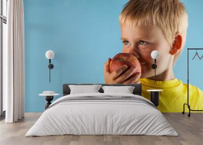 A pretty blond boy sniffs a red Apple and smiles against a blue background. The concept of healthy eating. Copy space. Wall mural