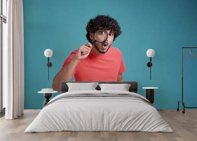 Funny Indian man looking through magnifying glass, searching or investigating something, standing in coral t-shirt on blue studio background Wall mural