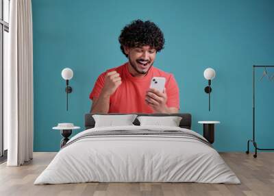 Happy indian man winner using cell mobile phone Excited ethnic guy holding smartphone feels surprised about betting win on cellphone, celebrating success in coral t-shirt on blue studio background Wall mural