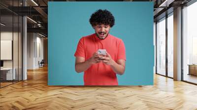 young latin man looks delighted into smartphone and says wow. Dressed in casual on a blue studio background Wall mural