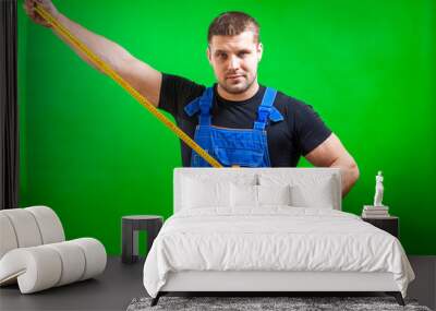 A dark-haired male builder in a black T-shirt and blue construction jumpsuit shows a long yellow tape rule on a green isolated background Wall mural