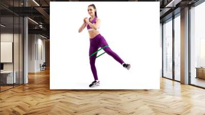 A dark-haired woman coach in a sporty purple  short top and gym leggings makes lunges  by the feet forward with sport fitness rubber bands,  stretch legs on a  white isolated background in studio Wall mural