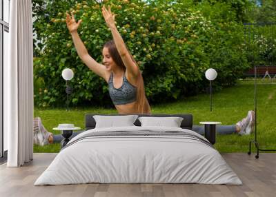 A dark-haired woman coach in a sporty short top and gym leggings makes a wide twine on the rug for yoga; hands are raised upwards on the yoga mat, hands are raised upwards,  on a summer day  Wall mural